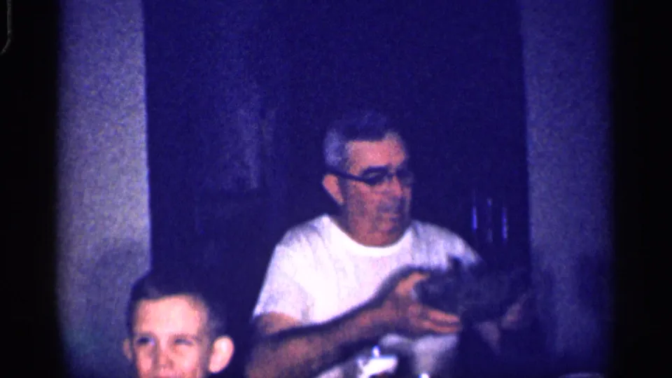 A man in a white shirt and glasses sits at a table in a dim 1959 Michigan home interior, with another adult partly visible.