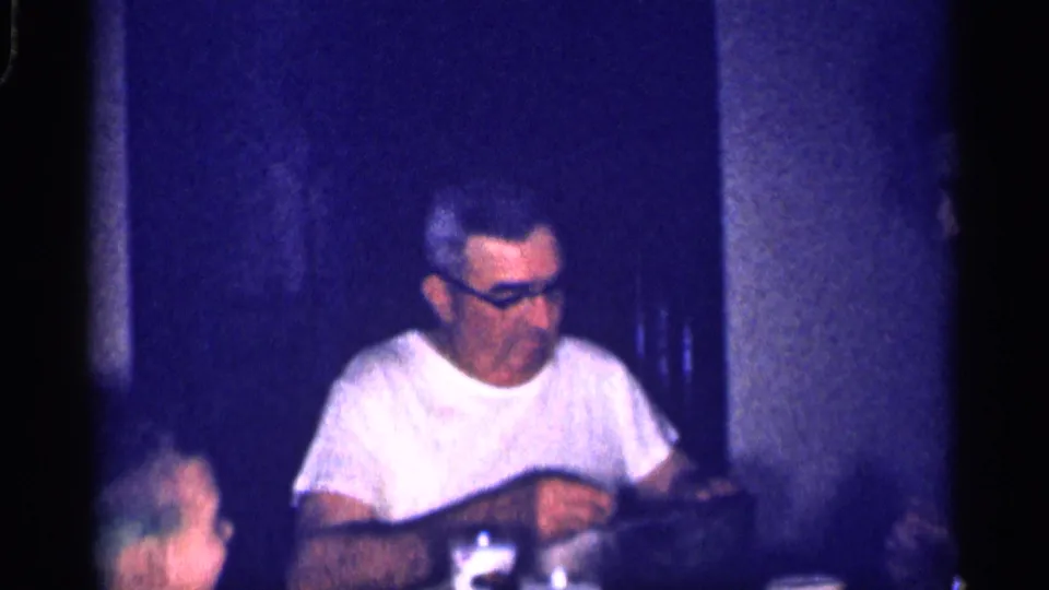 The man in a white shirt and glasses remains seated at the table as other family members are partly visible in the dim home interior.