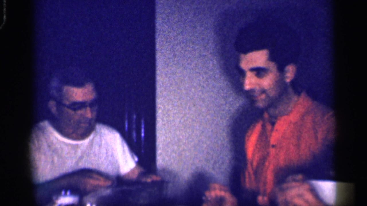 Two adult men seated at a table in a dim room, one in glasses and a white T-shirt and one in an orange shirt.