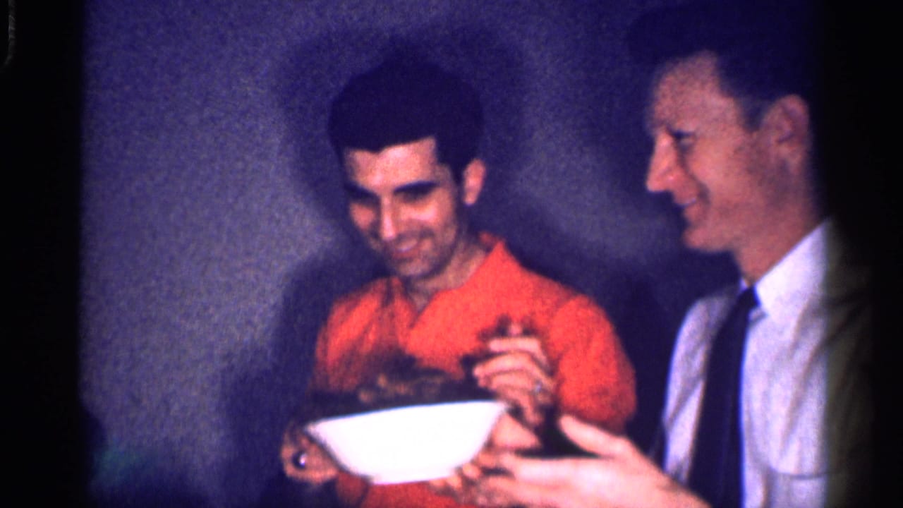 1959 Michigan frame of two adult men sharing food indoors, one holding a white bowl while the other smiles in a shirt and tie.