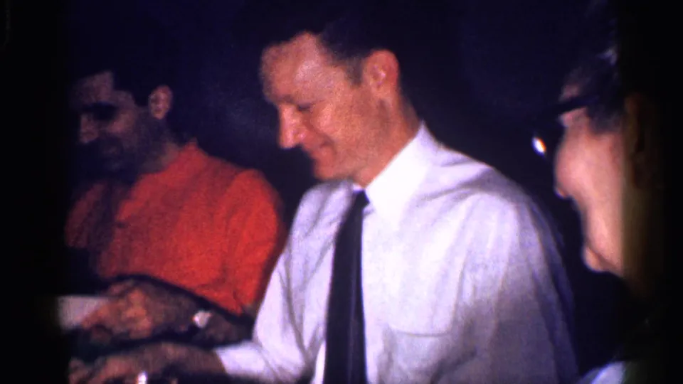 1959 Michigan home-movie frame showing adults gathered at a table, with food or dishes partly visible in a dark interior.