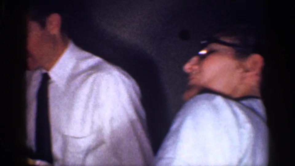 1959 Michigan frame showing a man in a white shirt and tie near a woman with glasses during an indoor home gathering.