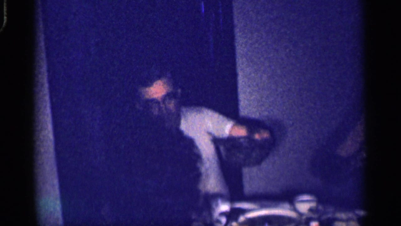 Man in a light shirt leans near the dinner table in a dark room.