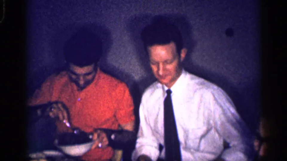 Two men sit at a dinner table in a 1959 Michigan home movie, one serving from a bowl beside a man in a tie.