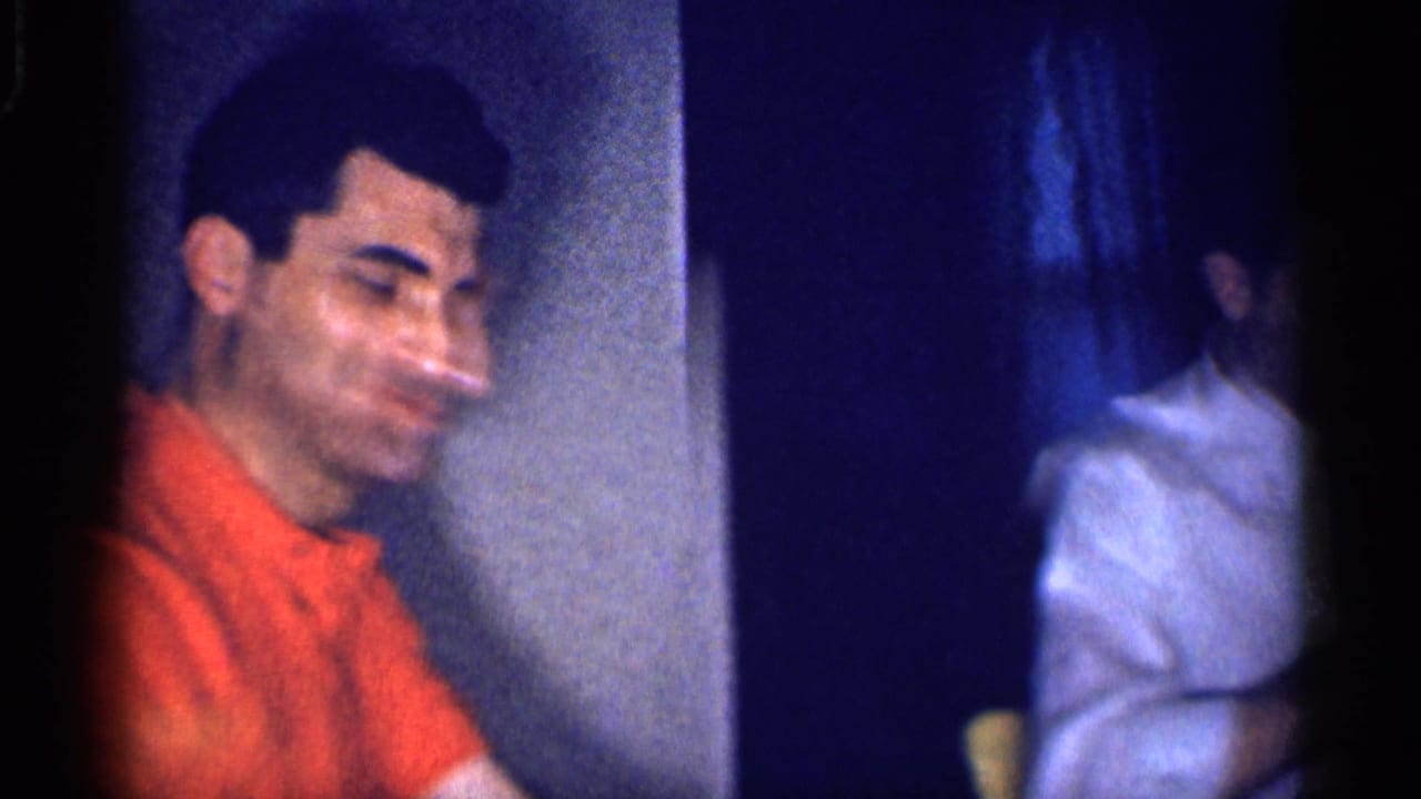 Man in an orange shirt smiles beside a dim dining table.