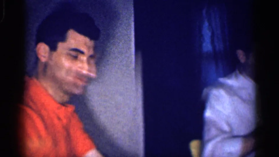 In a 1959 Michigan home-movie frame, an adult man in a bright orange shirt smiles near a table in a dim private interior.