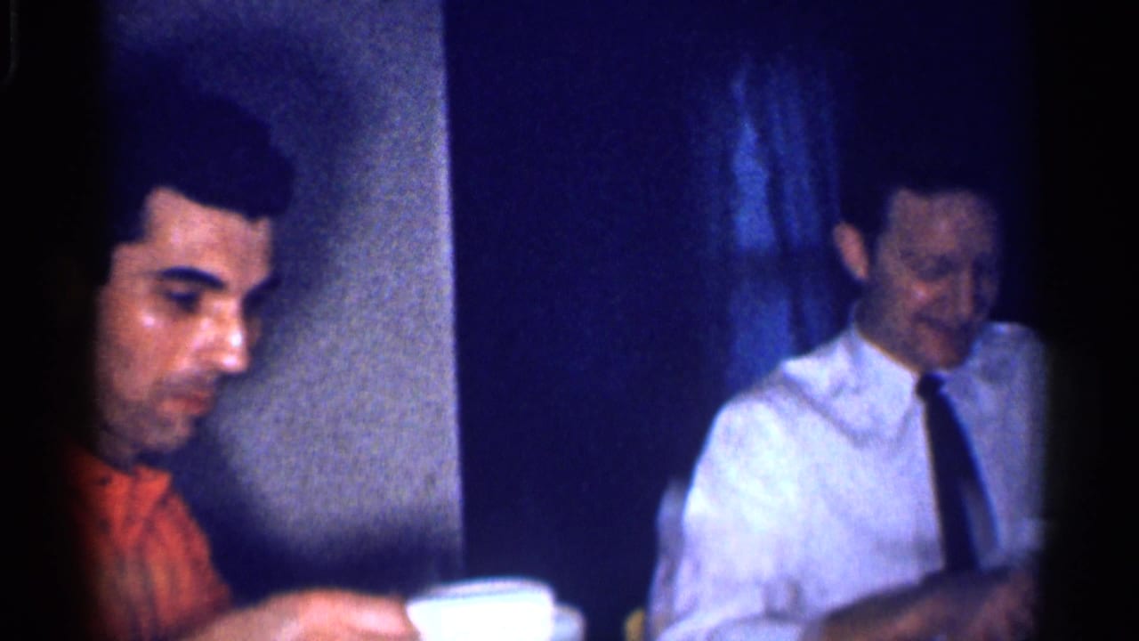 A 1959 Michigan interior frame shows two adult men at a table, with one in an orange shirt and another in a white shirt with a black tie.