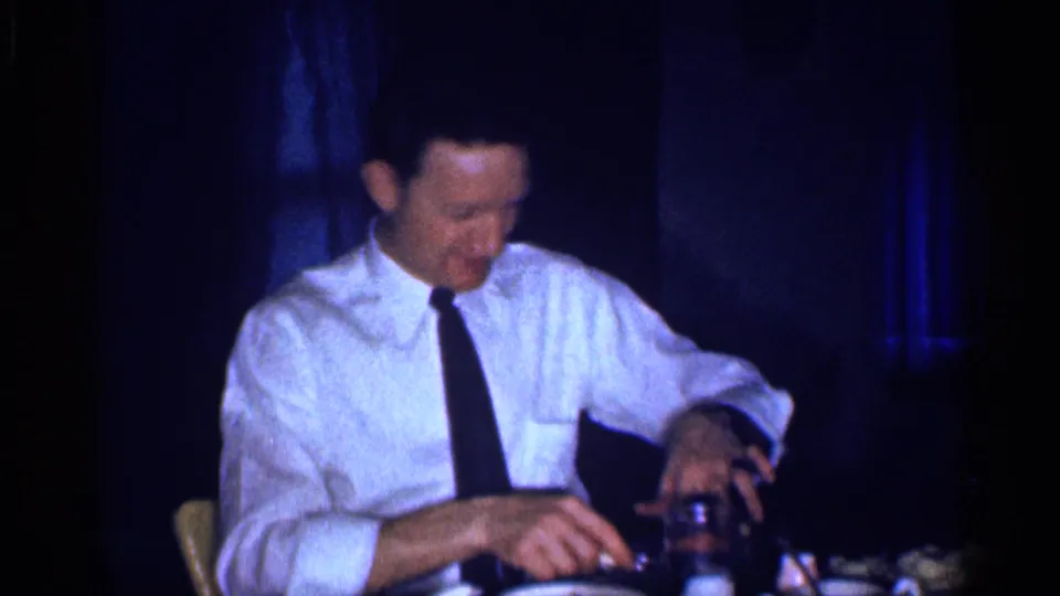 An adult man in a white dress shirt and black tie looks down while handling table items in a dim 1959 Michigan home-movie dinner scene.