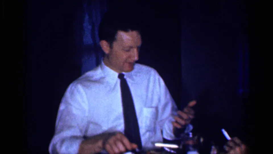 A man in a white shirt and black tie gestures with one hand while seated at a dinner table in 1959 Michigan home-movie footage.