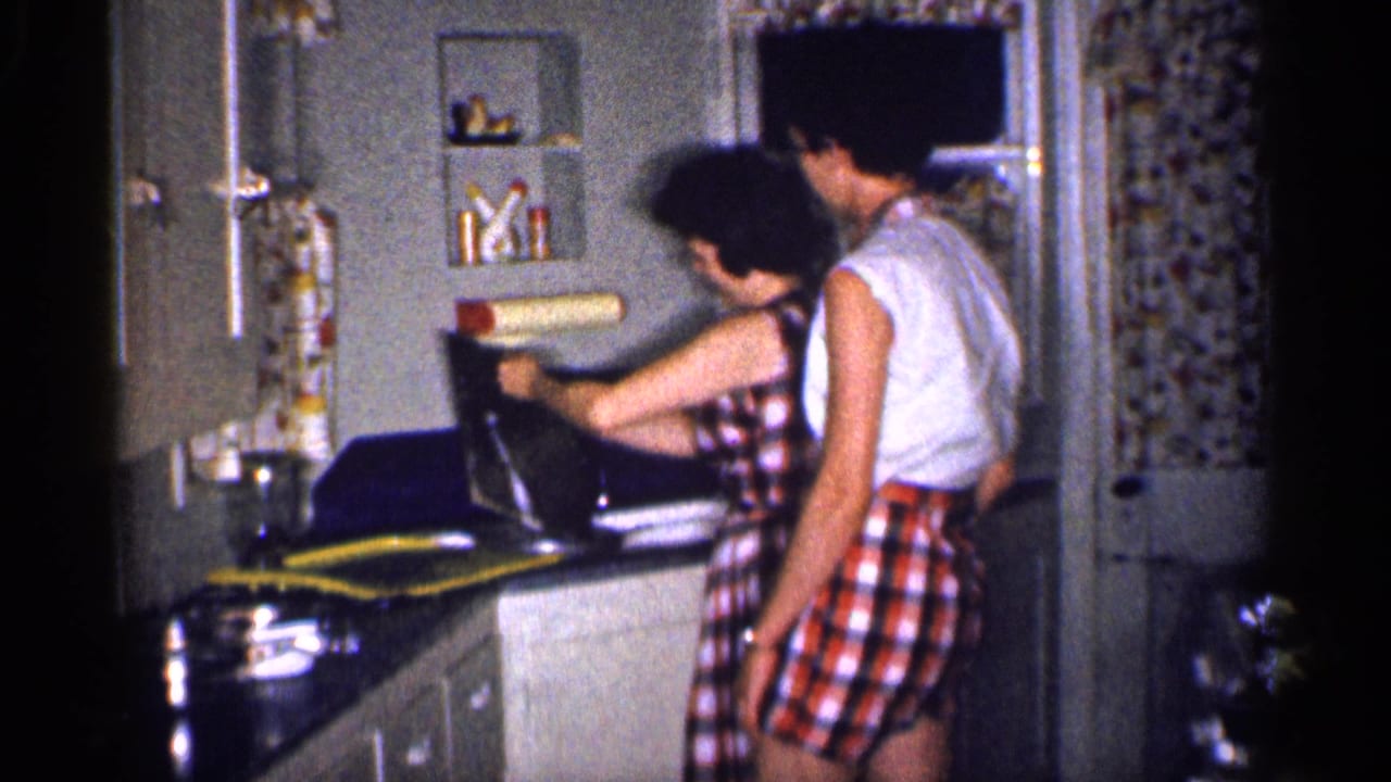 In a 1959 Michigan home kitchen, a woman in a checkered dress works at the sink while a man in a sleeveless shirt and checkered shorts stands close by.