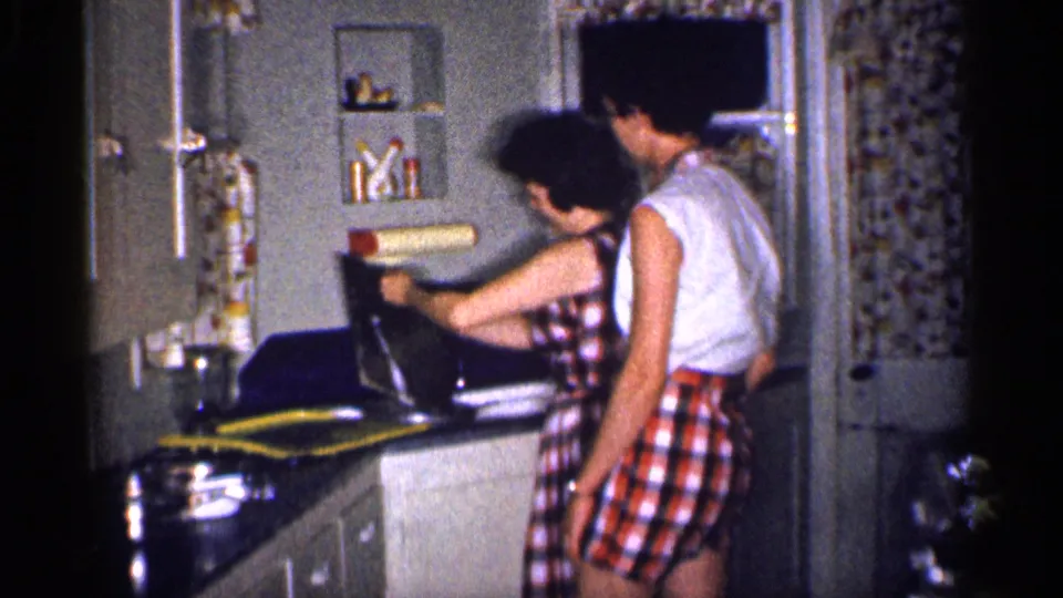 In a 1959 Michigan home kitchen, a woman in a checkered dress works at the sink while a man in a sleeveless shirt and checkered shorts stands close by.