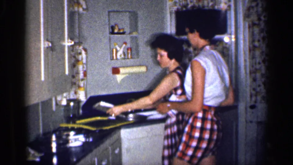 A dishwashing moment in the home kitchen shows the woman reaching across the sink area as the man stands nearby, with mid-century fixtures visible.