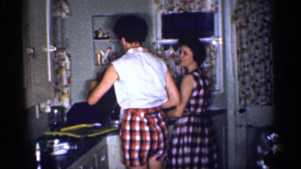 The man stands with his back to the camera at the sink while the woman turns toward him, framed by the kitchen window curtain and wall shelves.