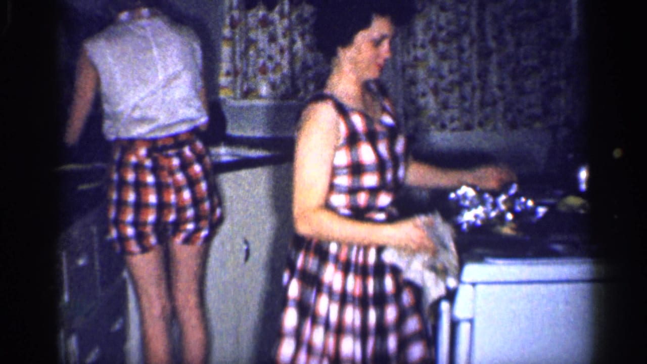 1959 Michigan kitchen frame showing a woman in a sleeveless plaid dress handling a cloth beside dishes.