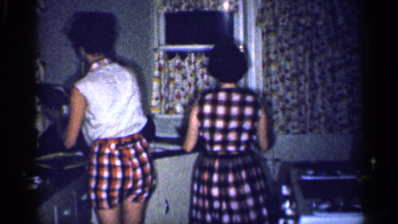 Home-movie frame from 1959 Michigan showing two people from behind at a kitchen counter below floral curtains.
