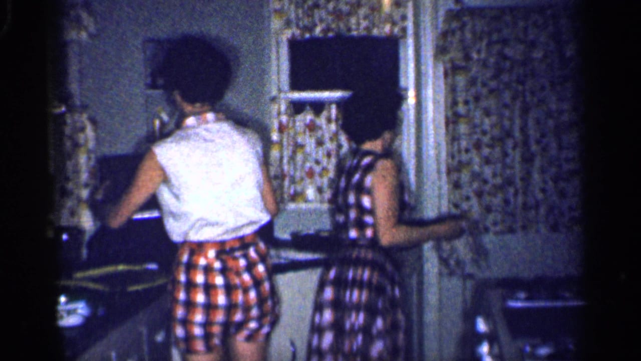 Two women stand at a kitchen counter near floral curtains and a small window.