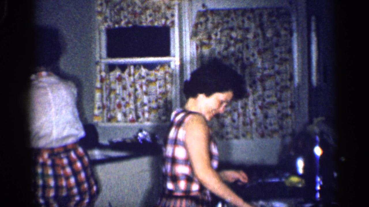 The women continue tidying in the 1959 Michigan kitchen, one looking down near the counter and one turned away.