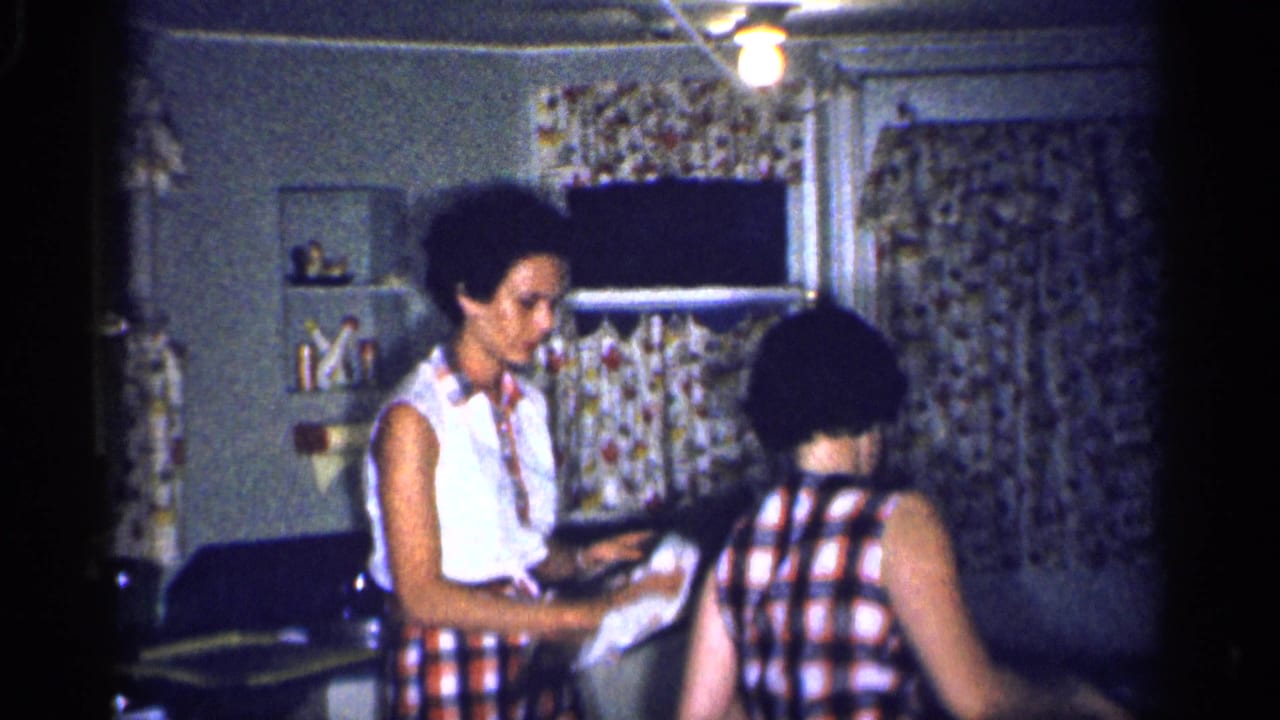 Two women tidy together at the counter in a modest 1959 Michigan kitchen with floral curtains and low light.