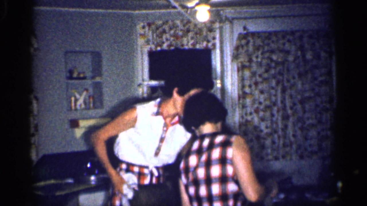 In a 1959 Michigan home kitchen, two adult women in summer clothing lean close while talking near a table and floral curtains.