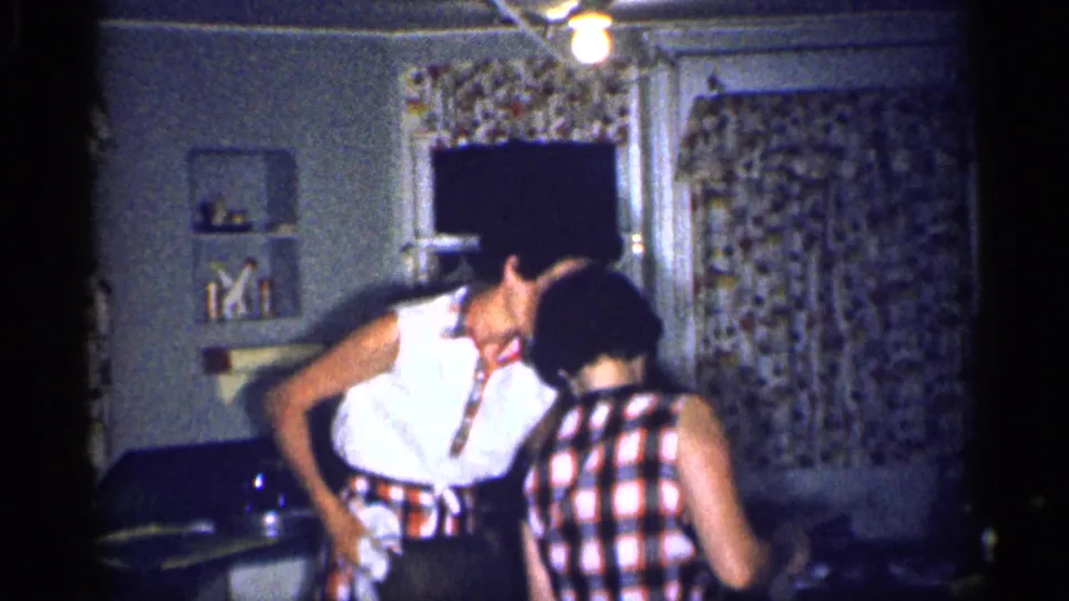 In a 1959 Michigan home kitchen, two adult women in summer clothing lean close while talking near a table and floral curtains.