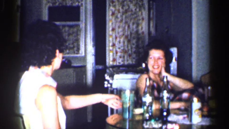 Two adult women sit at a table in a Michigan kitchen, with bottles and cups visible as one smiles toward the camera.