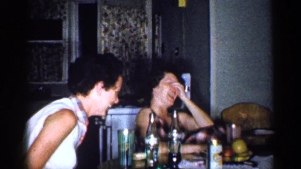 In the 1959 Michigan home interior, two women laugh across a kitchen table set with bottles, cups, and fruit.