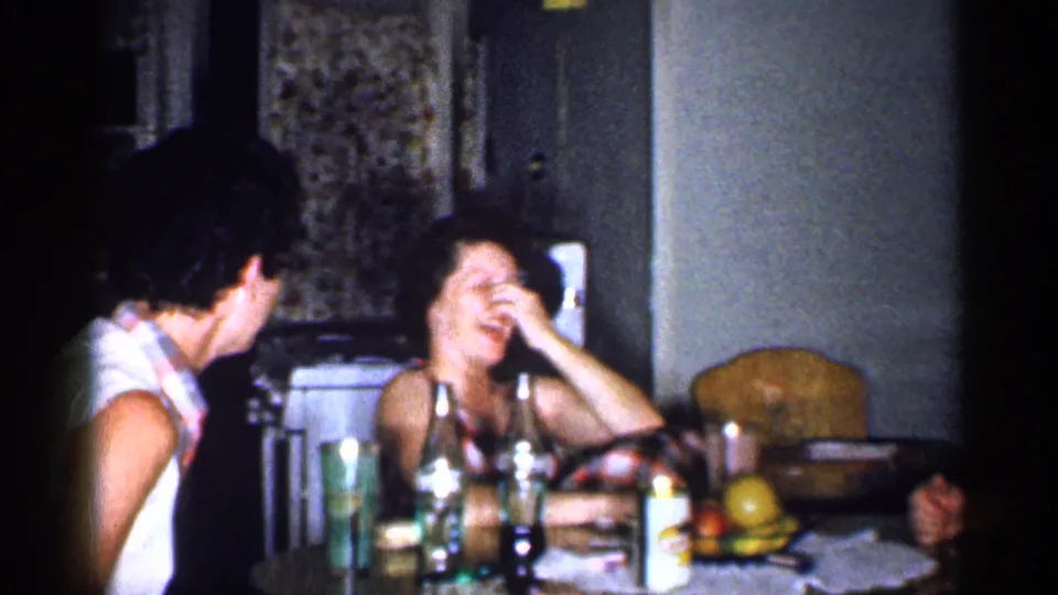 Two adult women continue laughing in a 1959 Michigan kitchen, with bottles, fruit, and chairs visible around the table.