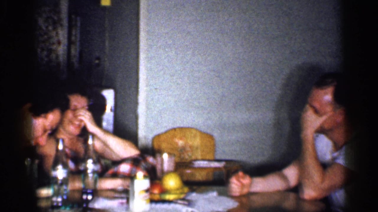 Three adults sit around a kitchen table with bottles and fruit in a dim interior.