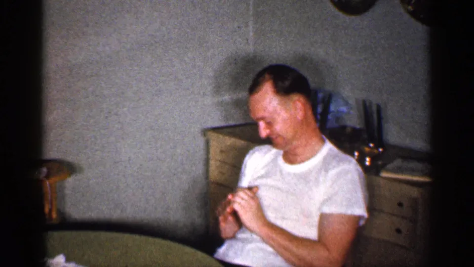 A man in a white t-shirt sits at a kitchen table in a 1959 Michigan home interior, with a cabinet and household objects behind him.