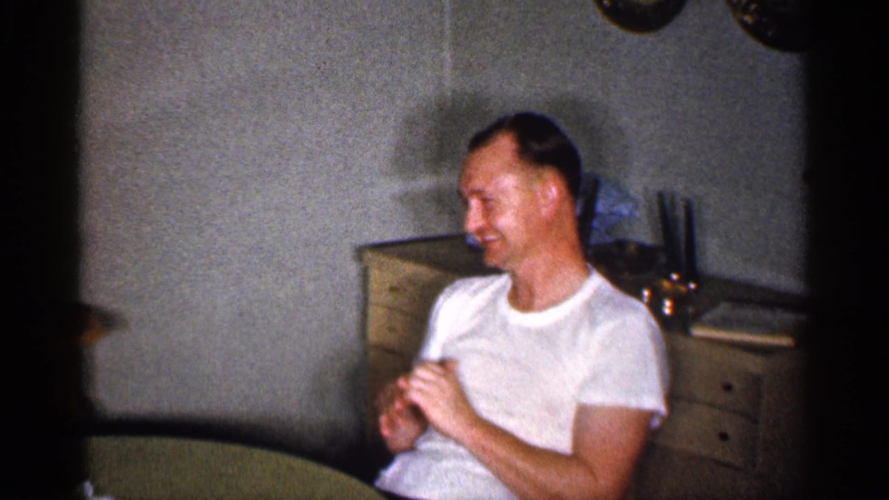 Man in a white T-shirt sits smiling at a round table in a home interior.