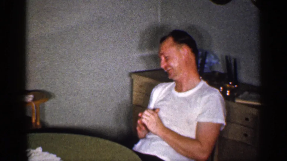 A man seated in a private Michigan interior smiles with clasped hands near a round table, captured on 8mm film in 1959.