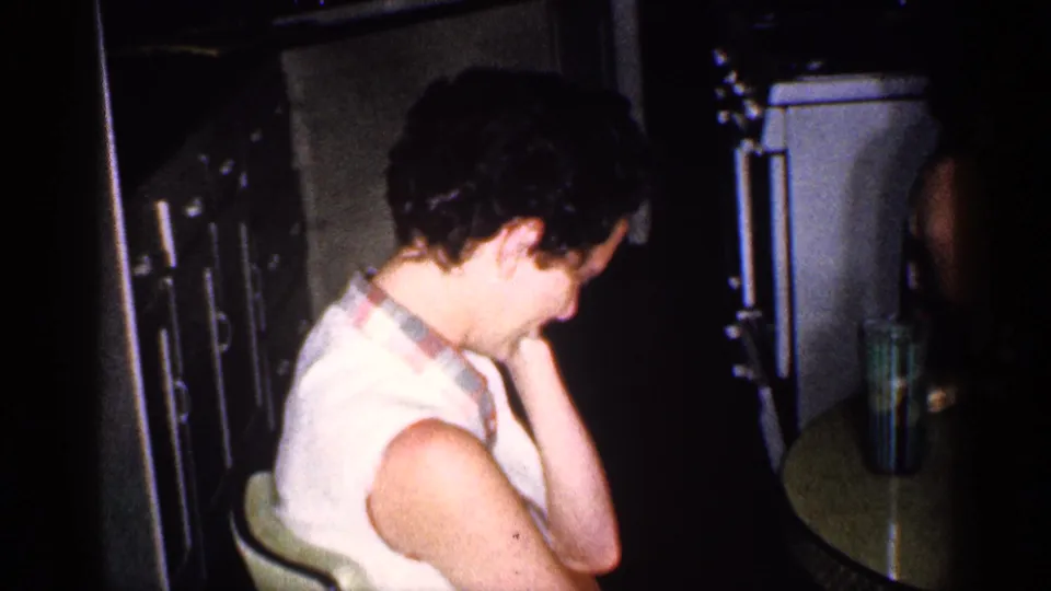 A woman in a sleeveless top sits near a round table in the same 1959 Michigan private interior, looking downward or aside.