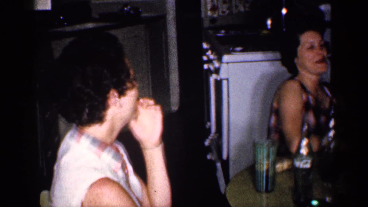 Two adults sit at a kitchen table with glasses and bottles nearby.