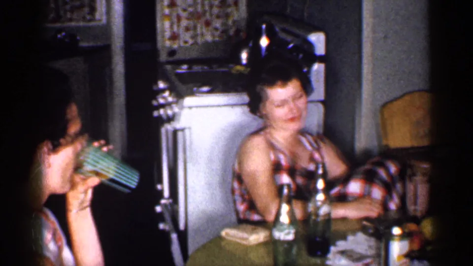 1959 Michigan home-movie frame showing a woman sipping from a striped green glass as another woman sits beside bottles in a kitchen.