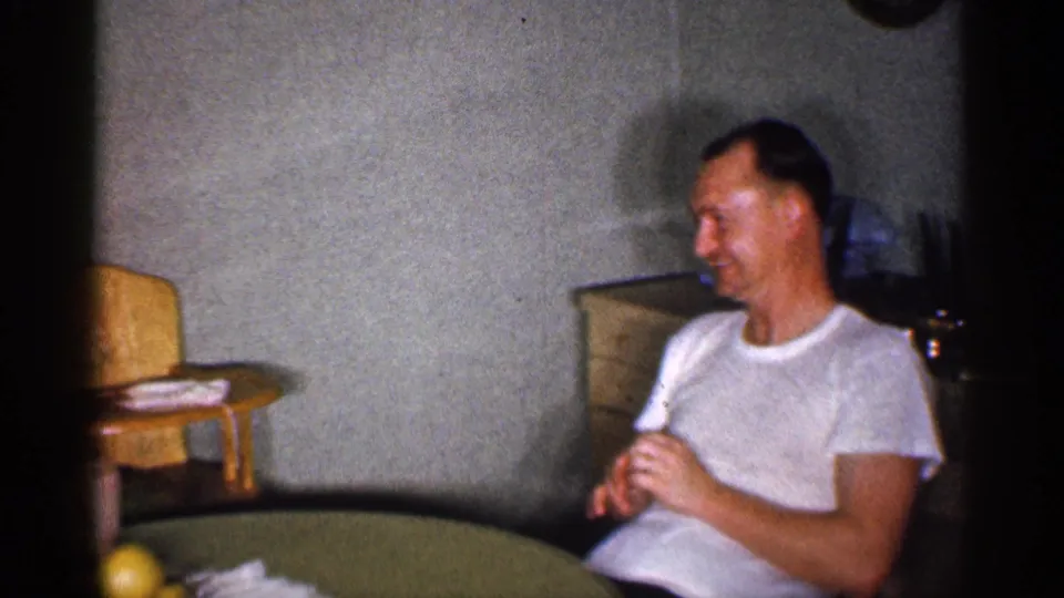 1959 Michigan home-movie frame of a man in a white T-shirt seated beside a round kitchen table in a private interior.