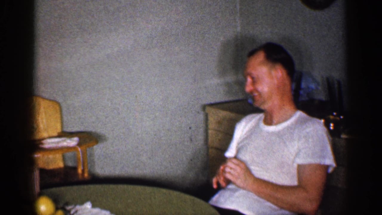Adult man in a white T-shirt sitting beside a small table in a living room.