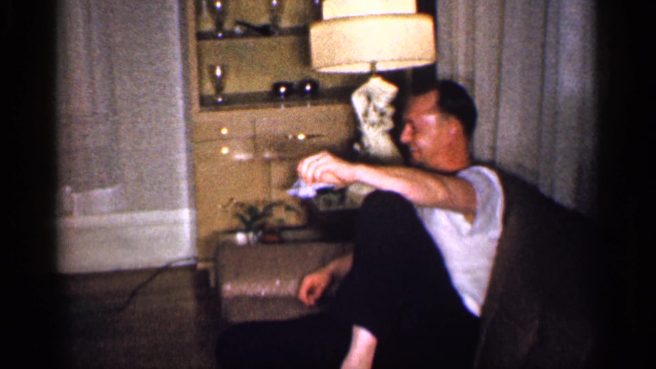 An adult man sits in a brown chair in a Michigan living room, with a lit tan lamp, cabinet, and curtains behind him.