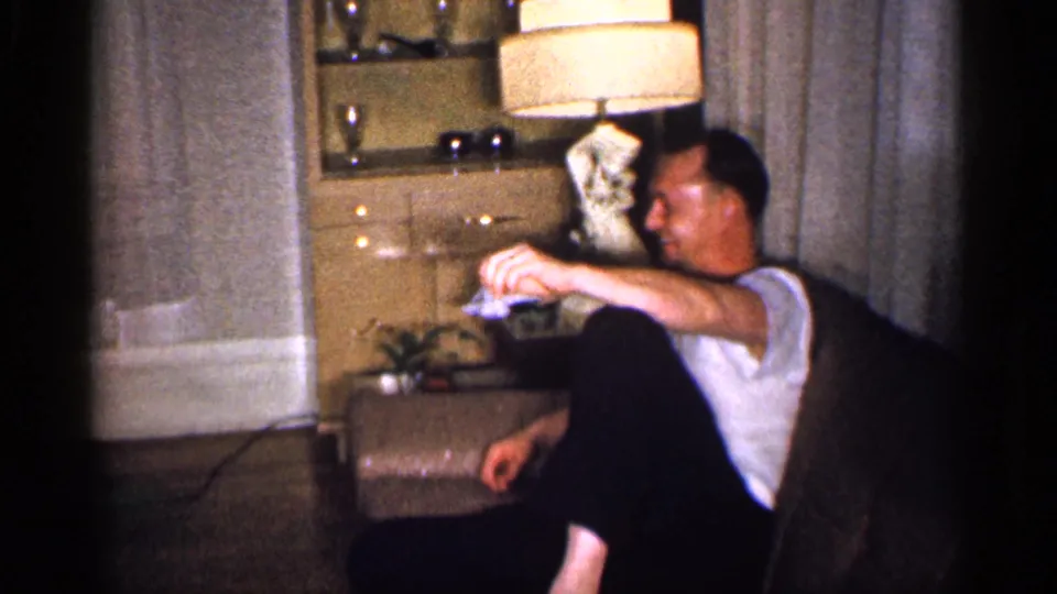 An adult man sits in a brown chair in a Michigan living room, with a lit tan lamp, cabinet, and curtains behind him.