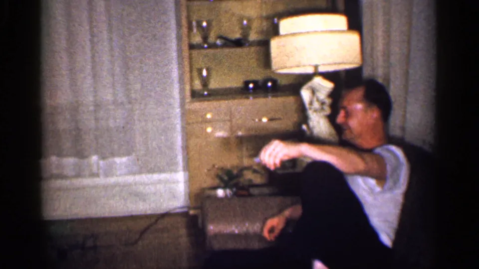 The man remains seated in the living room, leaning back near a tan lamp, cabinet, curtained window, and small plant.