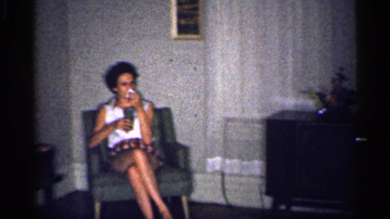 Woman seated in an armchair, holding a glass in a living room with curtains and a cabinet nearby.