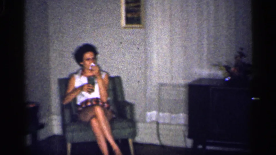 1959 Michigan home-movie frame of a woman relaxing in a green armchair inside a private living room, holding a drink.