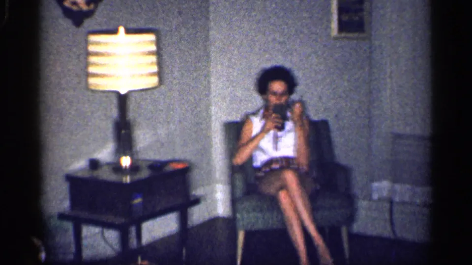 1959 Michigan home-movie frame showing a woman in an armchair beside a lit striped lamp, side table, and curtained wall.