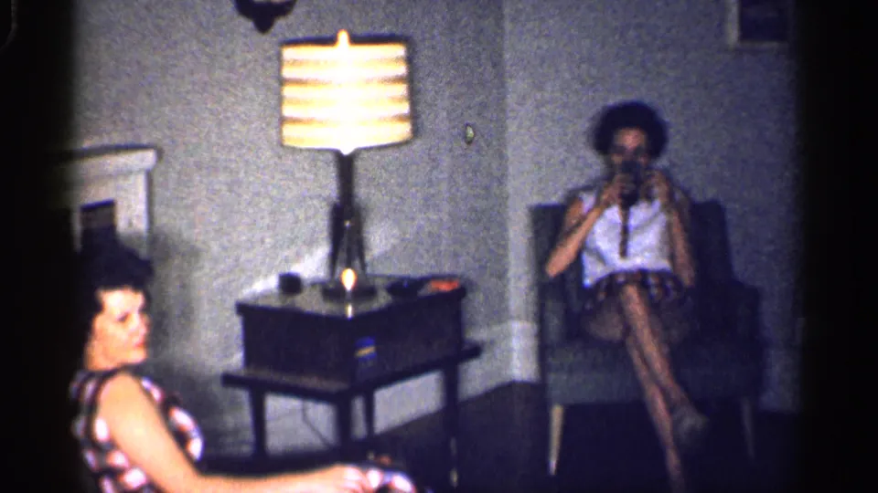 1959 Michigan home-movie frame with two women in a private living room, a lit striped lamp, side table, and armchair.