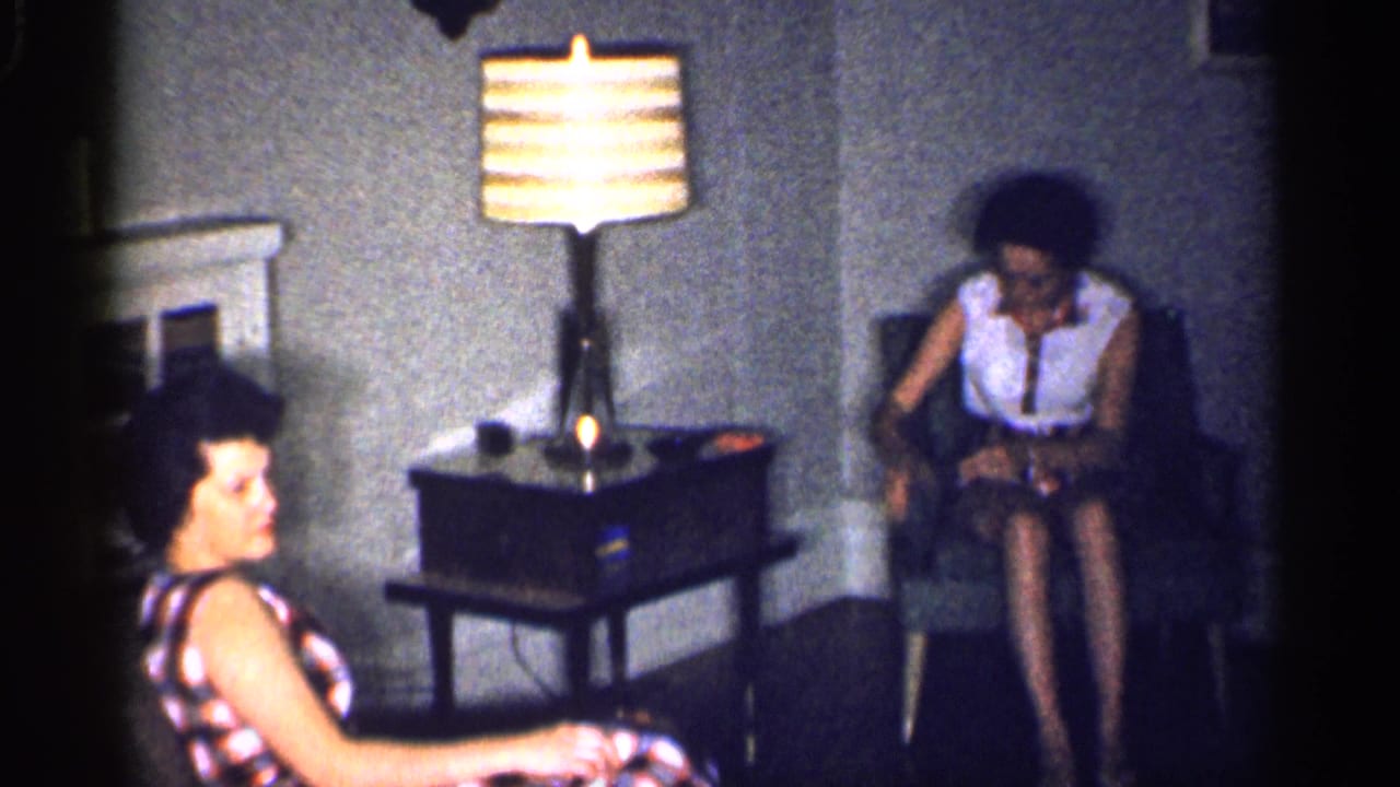 Two women sit in a dim living room beside a lit table lamp and radio.