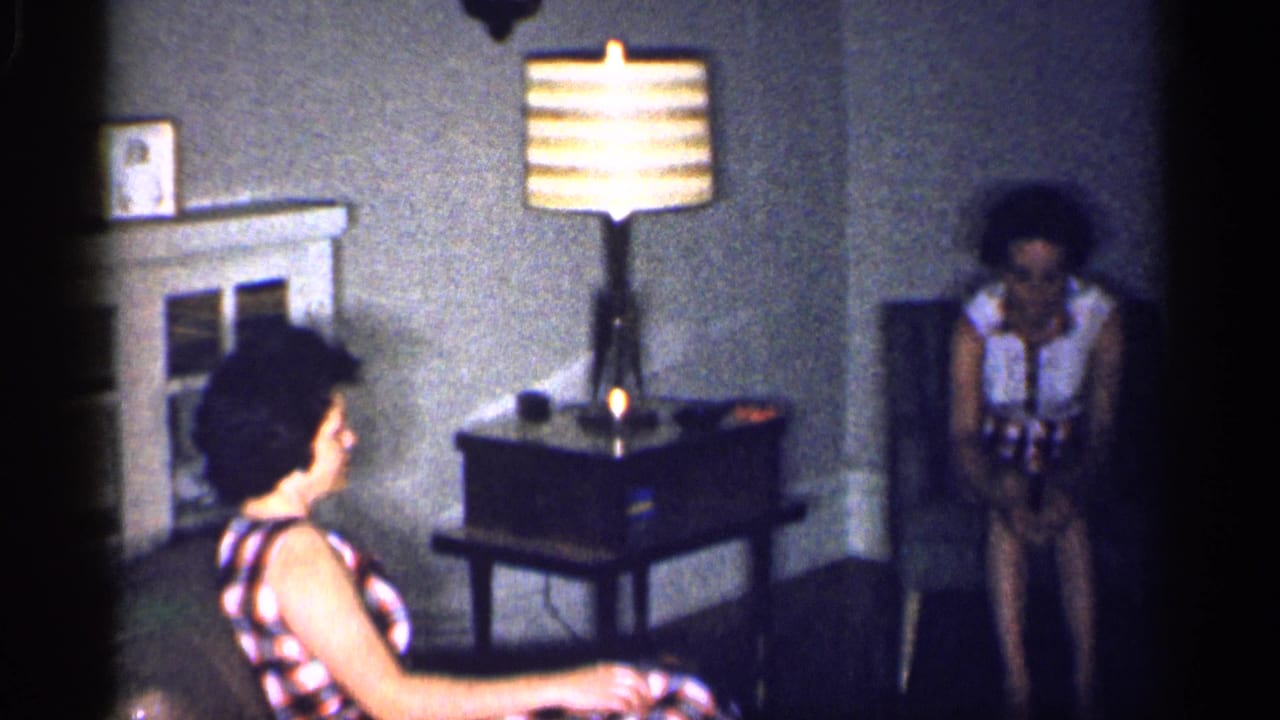 A woman in a plaid dress sits in profile while another woman sits near the corner table in a 1959 Michigan living room.