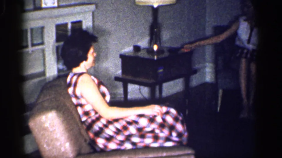 In the living room, the seated woman in plaid looks toward another woman reaching near the table with the lamp and radio.