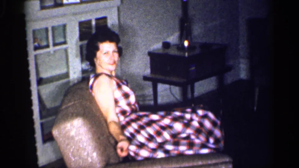 A woman in a plaid dress smiles from an upholstered chair beside a lamp-lit table in the Michigan home interior.
