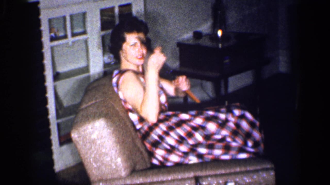 In a 1959 Michigan home-movie frame, a woman in a plaid dress sits near a fireplace and holds a toy gun toward the camera.