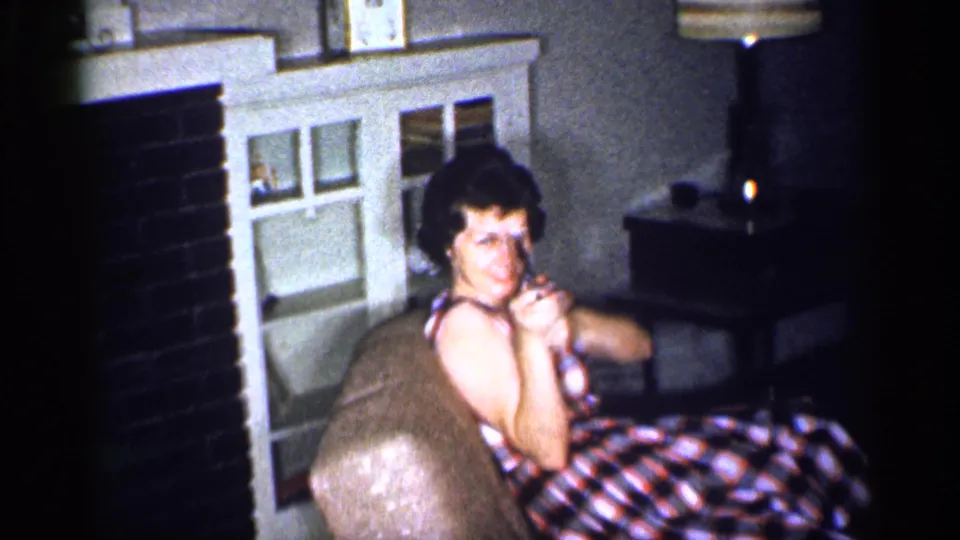 A woman in a plaid dress sits beside a fireplace in a Michigan living room, playfully aiming a toy gun during home-movie footage.