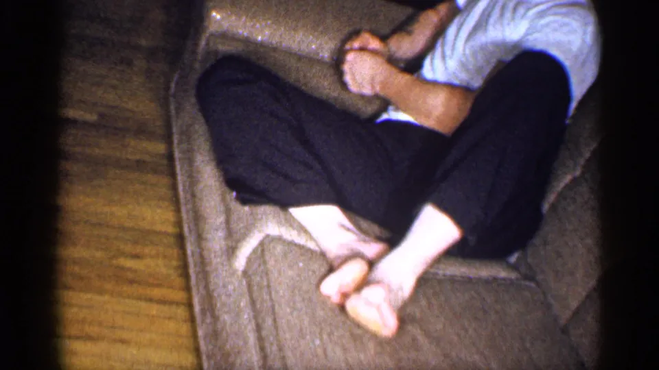 The camera frames a seated adult’s crossed legs and bare feet on an upholstered chair, with a wood floor visible in the home interior.